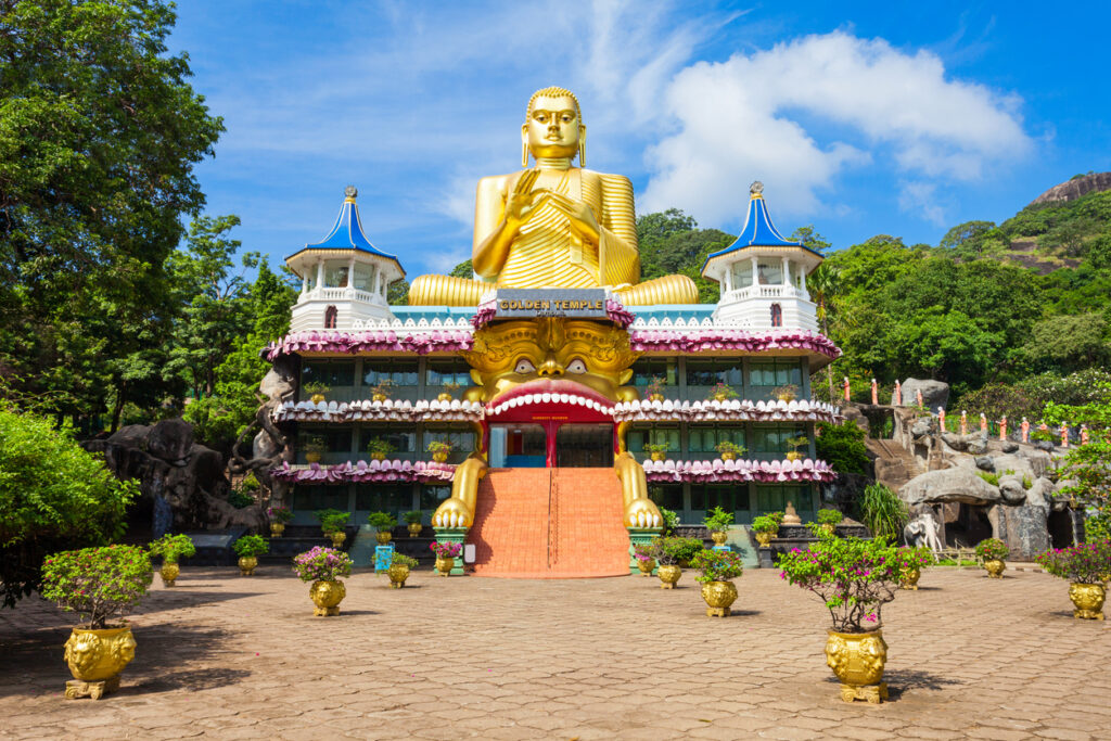 Frontansicht des Golden Temple in Dambulla mit monumentaler sitzender goldener Buddhafigur, blauweißen Türmen und Löwenmaul-Eingang unter blauem Himmel.