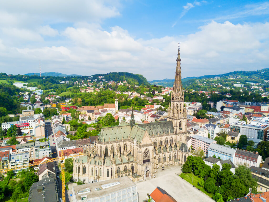 Luftaufnahme des Linzer Mariendoms mit spitzem Turm, umgeben von Stadtvierteln und grünen Hügeln.
