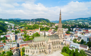 Luftaufnahme des Linzer Mariendoms mit spitzem Turm, umgeben von Stadtvierteln und grünen Hügeln.