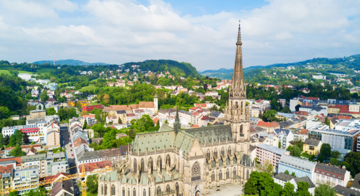 Luftaufnahme des Linzer Mariendoms mit spitzem Turm, umgeben von Stadtvierteln und grünen Hügeln.