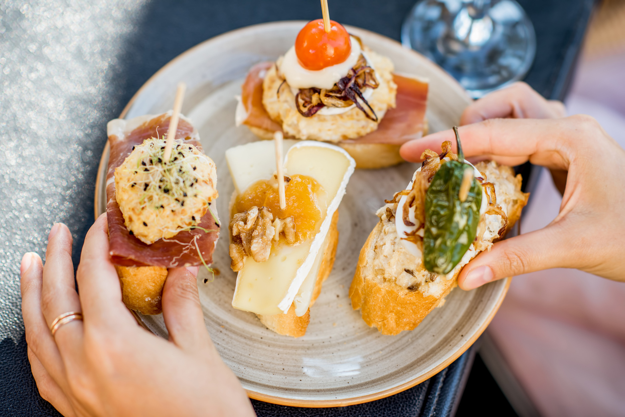 Teller mit vier baskischen Pintxos auf Baguette – Schinken mit Creme, Käse mit Walnuss und Chutney, Thunfischsalat mit Pimiento sowie Ei-Mayonnaise mit Röstzwiebeln – von zwei Händen gehalten.