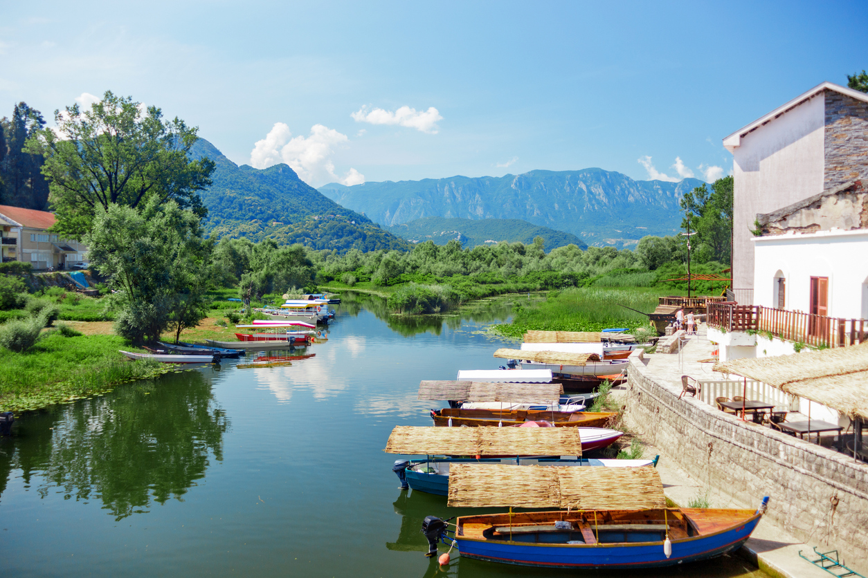 Kanal am Skadarsee mit hölzernen Ausflugsbooten mit Schilfdächern, Uferpromenade und grüner Bergkulisse.
