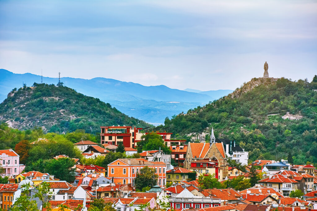 Panorama von Plovdiv mit roten Dächern, zwei bewaldeten Hügeln und dem Alyosha-Denkmal vor blauen Bergen.