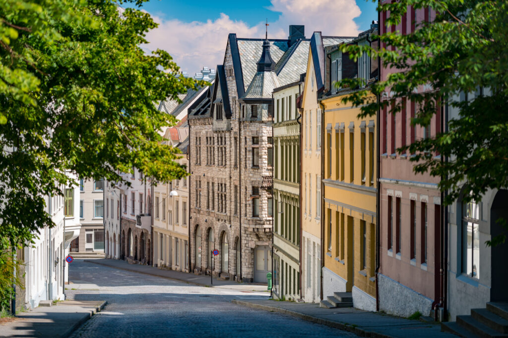 Baumgerahmte Kopfsteinpflasterstraße mit farbigen Jugendstilfassaden in Ålesund, Norwegen, unter blauem Himmel.