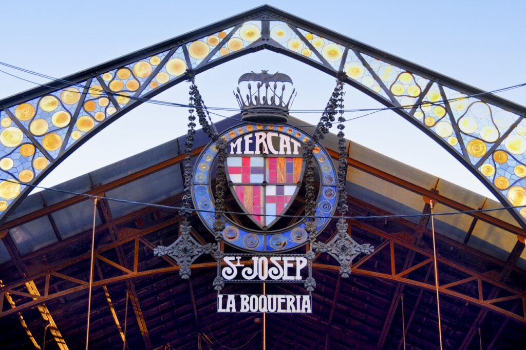 Detail des Glas- und Eisenportals mit Wappen und der Aufschrift „Mercat St Josep La Boqueria“ vor blauem Himmel.