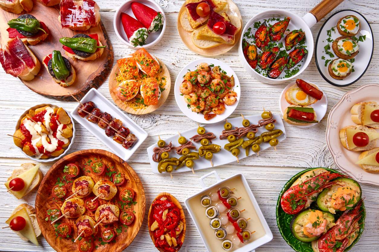 Draufsicht auf viele spanische Tapas und baskische Pintxos auf weißem Holztisch mit Oliven-Spießen, Gambas, Patatas Bravas, Pimientos, Jamón und Brot.
