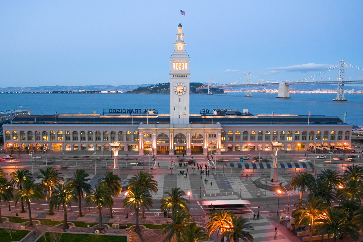Abendaufnahme des Ferry Building mit beleuchtetem Uhrenturm an der Embarcadero, dahinter die Bay Bridge und die Bucht von San Francisco.