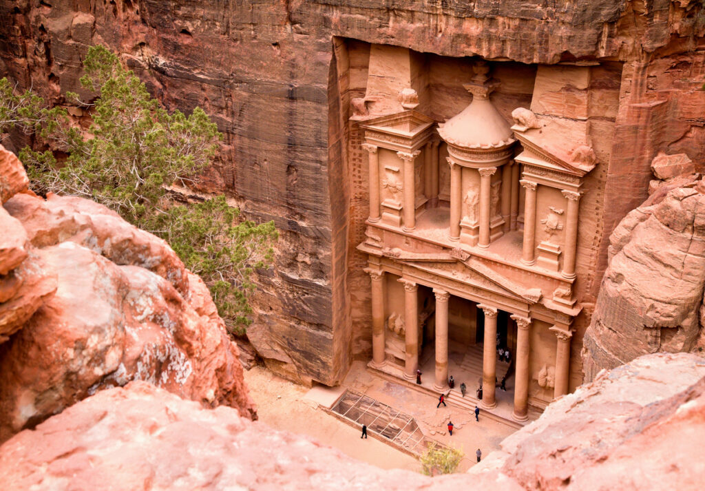 Obere Ansicht der rosafarbenen Felsfassade Al-Khazneh in Petra mit Menschen am Eingang.