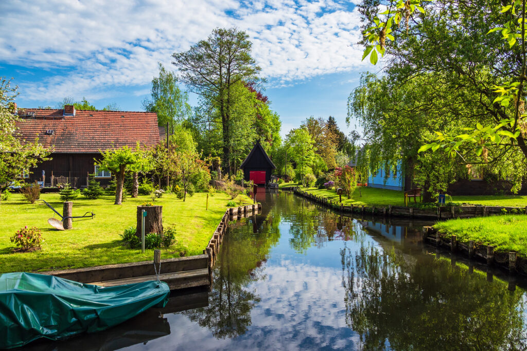Ein Klarwasserkanal spiegelt Gärten, Bäume und ein schwarzes Bootshaus mit roter Tür.