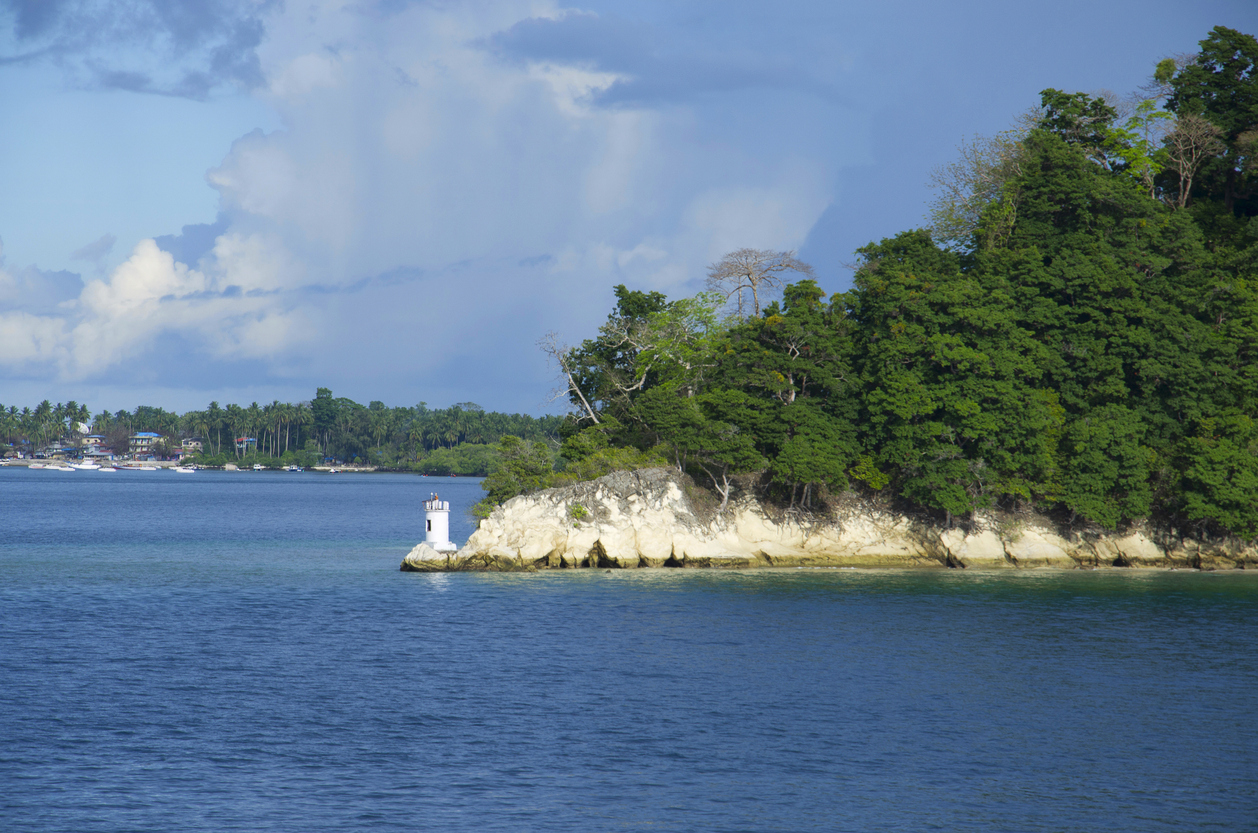 Blick auf eine bewaldete Landzunge bei Port Blair mit weißem Mini-Leuchtturm auf hellen Felsen und ruhigem, blauem Meer.
