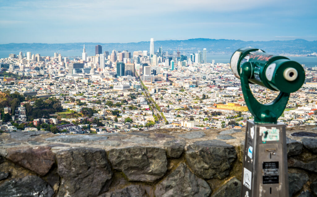 Blick von Twin Peaks über San Franciscos Skyline mit Market-Street-Achse und Bay, vorn ein grünes Aussichtsfernrohr an der Steinmauer.
