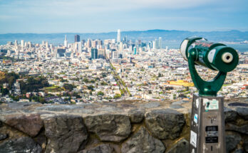 Blick von Twin Peaks über San Franciscos Skyline mit Market-Street-Achse und Bay, vorn ein grünes Aussichtsfernrohr an der Steinmauer.