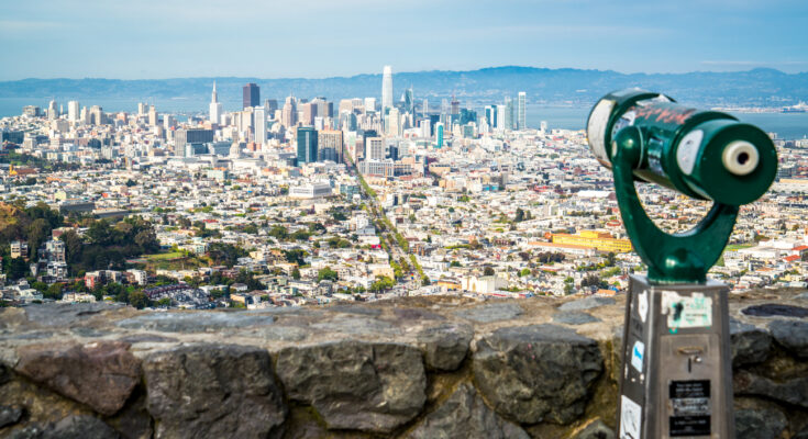 Blick von Twin Peaks über San Franciscos Skyline mit Market-Street-Achse und Bay, vorn ein grünes Aussichtsfernrohr an der Steinmauer.