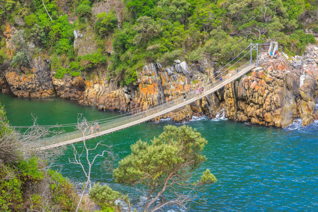 Schmale Fußgängerbrücke über türkisgrüner Flussmündung zwischen steilen Felsen und Küstenwald.