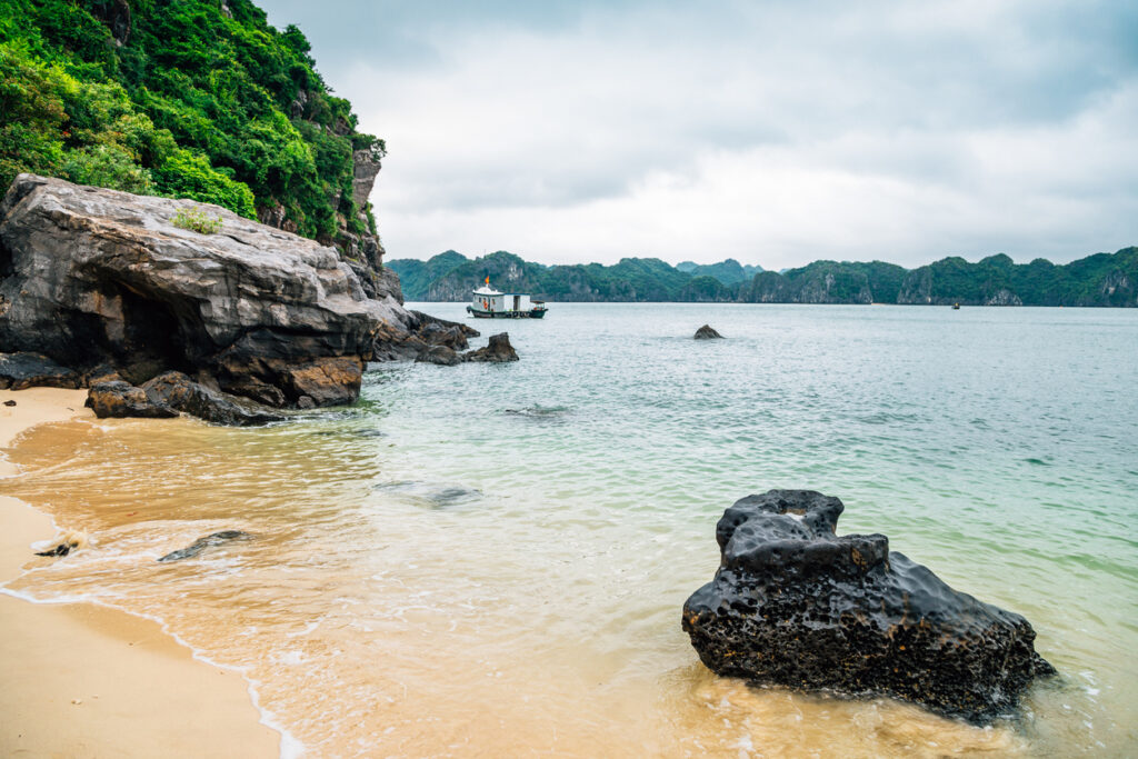 Kleiner Sandstrand mit Felsen, ruhigem Wasser und Fischerboot in der Ferne.