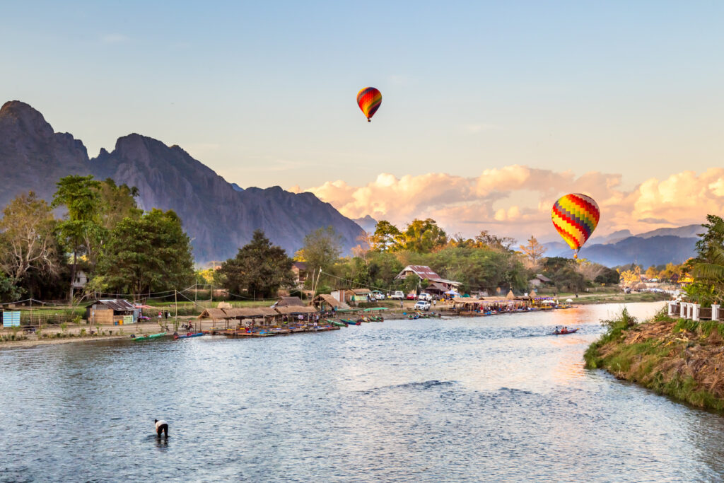 Bunte Ballons schweben bei Sonnenuntergang über Vang Vieng mit Karstbergen und Fluss.