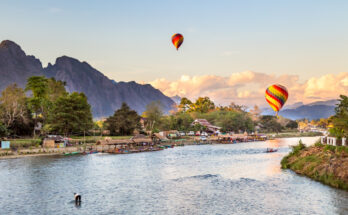 Bunte Ballons schweben bei Sonnenuntergang über Vang Vieng mit Karstbergen und Fluss.