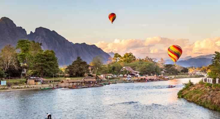 Bunte Ballons schweben bei Sonnenuntergang über Vang Vieng mit Karstbergen und Fluss.