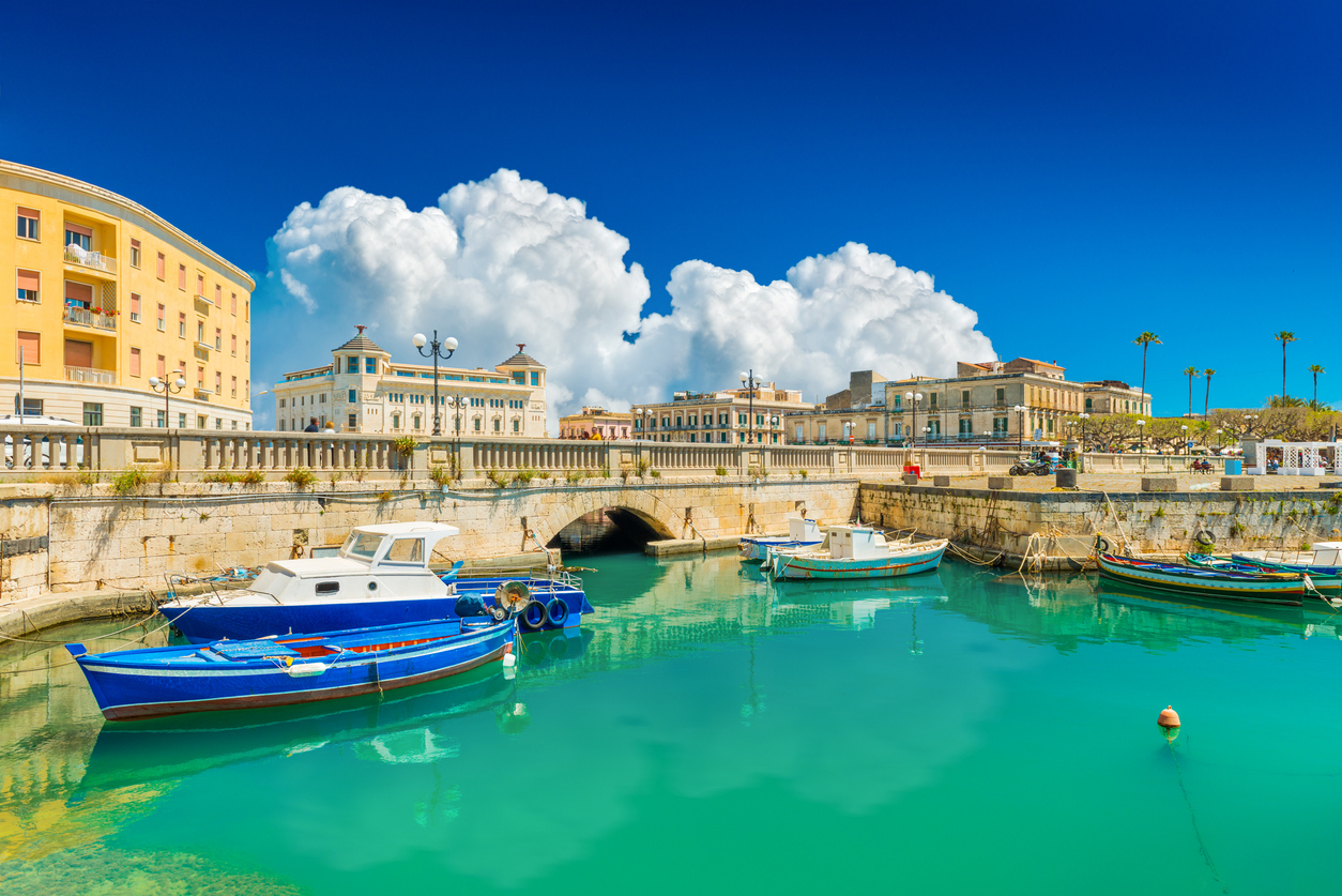 Historischer Hafen von Syrakus mit blau-weißen Fischerbooten, Steinbrücke und Palazzi unter tiefblauem Himmel.