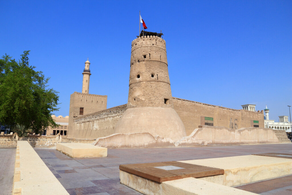 Sandfarbene Festung mit Rundturm und Flagge vor blauem Himmel.