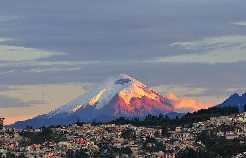 Schnee­bedeckter Vulkan hinter der Stadt Quito bei Sonnenuntergang.