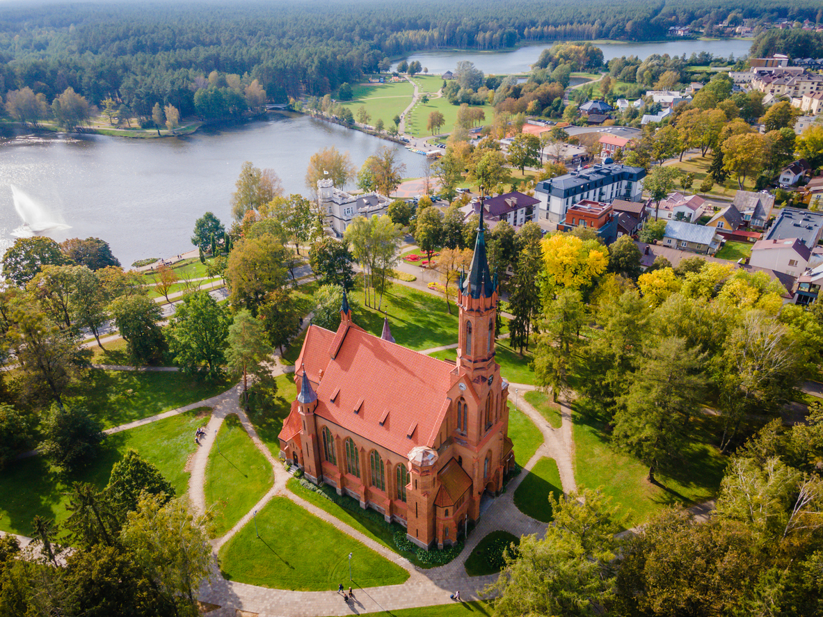 Luftaufnahme einer roten Kirche mit hohem Turm zwischen Bäumen und Seen.