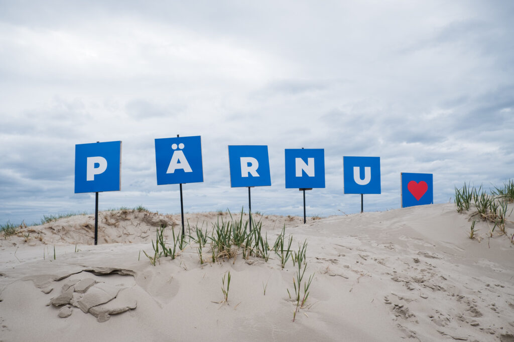 Blaue Tafeln mit der Aufschrift „PÄRNU“ stehen in sandigen Dünen am Ostseestrand.