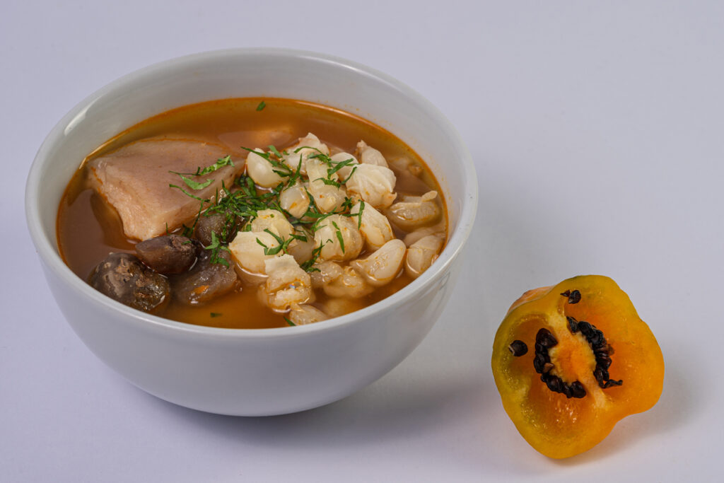 Weiße Schüssel mit kräftiger Suppe, Maiskörnern, Fleisch und halber gelber Paprika daneben.