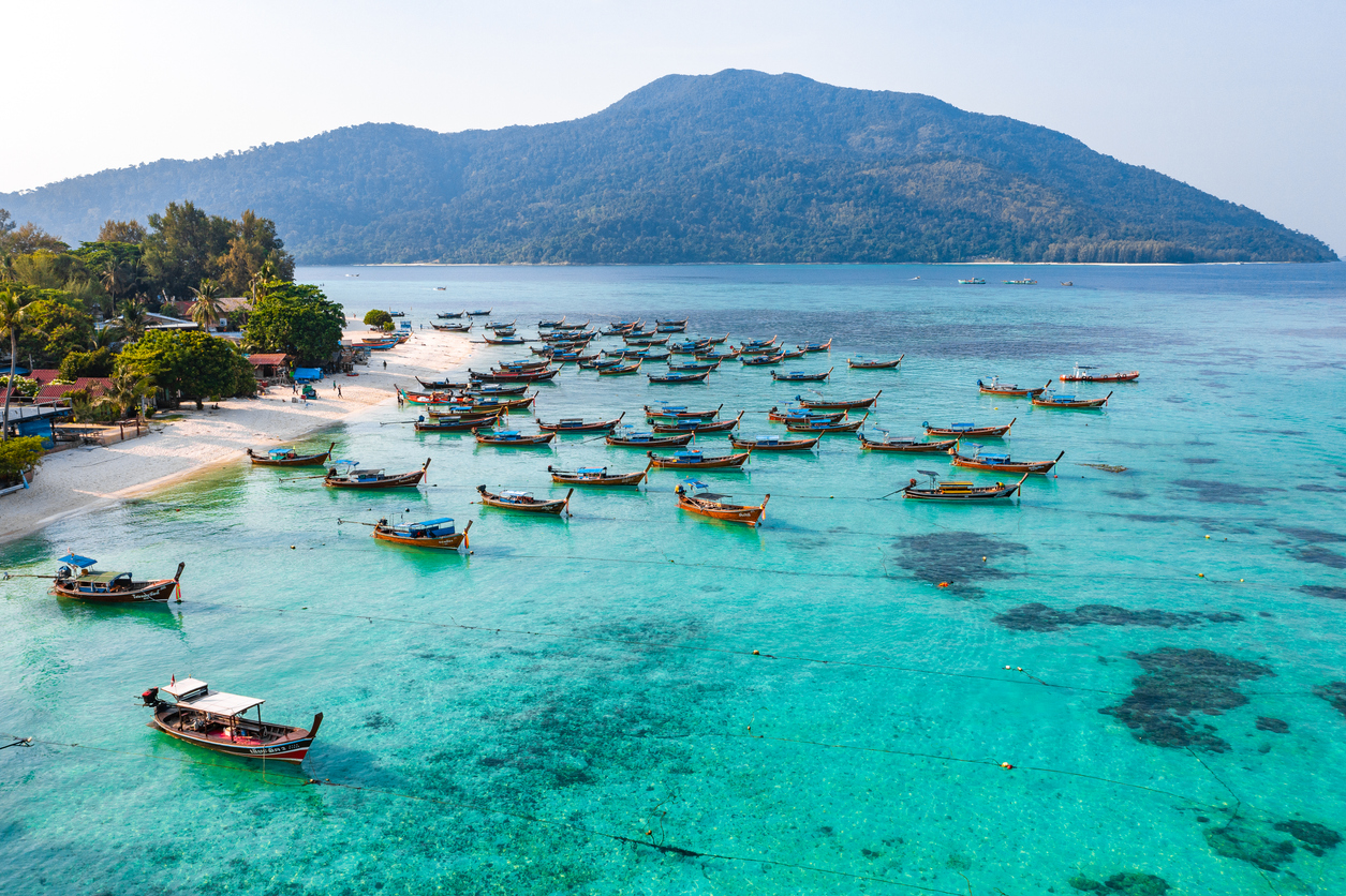 Viele Longtail-Boote liegen im türkisblauen Wasser an einem weißen Strand mit bewaldeter Insel im Hintergrund.