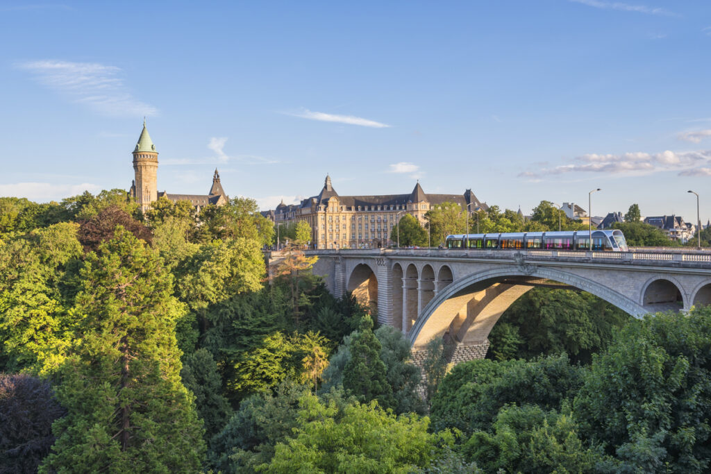 Straßenbahn fährt über eine Steinbogenbrücke vor einem Turm und vielen Bäumen.