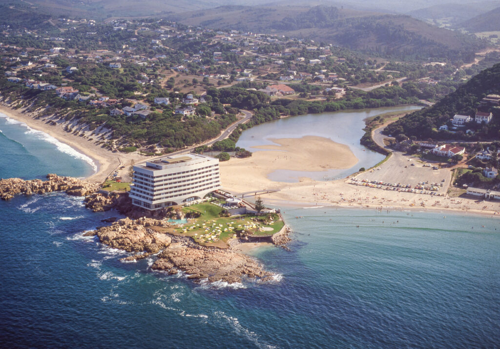 Luftaufnahme mit Hotel auf Felsen, breitem Sandstrand und ruhiger Lagune in Plettenberg Bay.