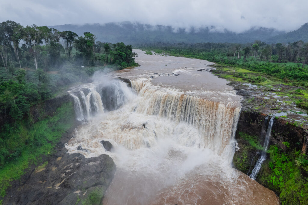Breiter Wasserfall auf dem Bolaven-Plateau mit braunem Hochwasser, Sprühnebel und grünem Regenwald.