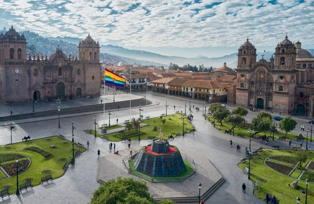 Blick über die Plaza de Armas in Cusco mit Kathedrale, Iglesia de la Compañía, grünen Beeten und der Regenbogen-Flagge.