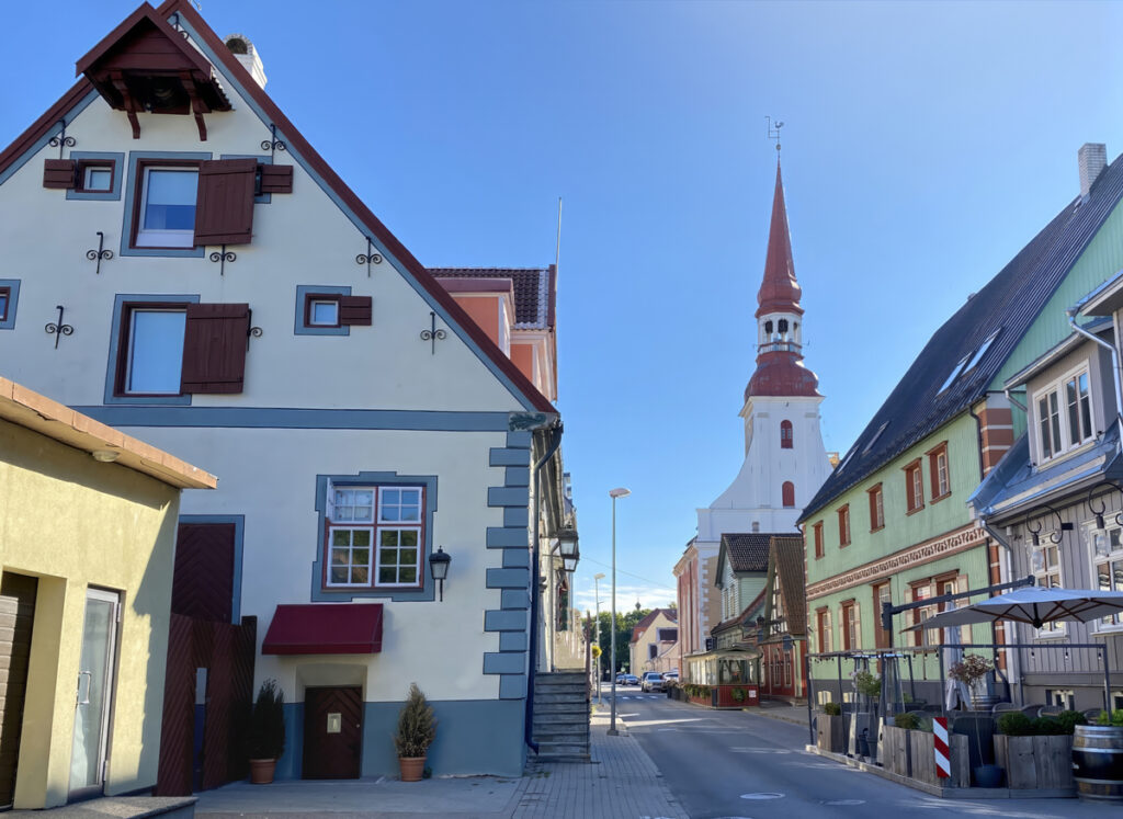 Bunte Holzhäuser und ein roter Kirchturm stehen an einer sonnigen Straße in Pärnu.