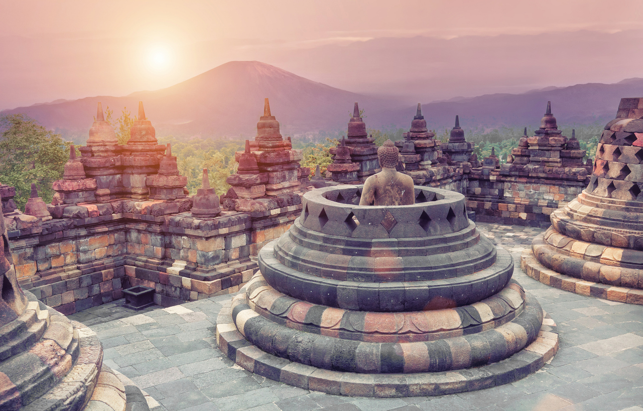 Buddhafigur im Borobudur-Tempel mit Stupas und Vulkan im rosa Morgenlicht.