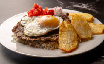 Teller mit Silpancho aus Reis, paniertem Fleisch, Spiegelei, Kartoffeln und Gemüse.