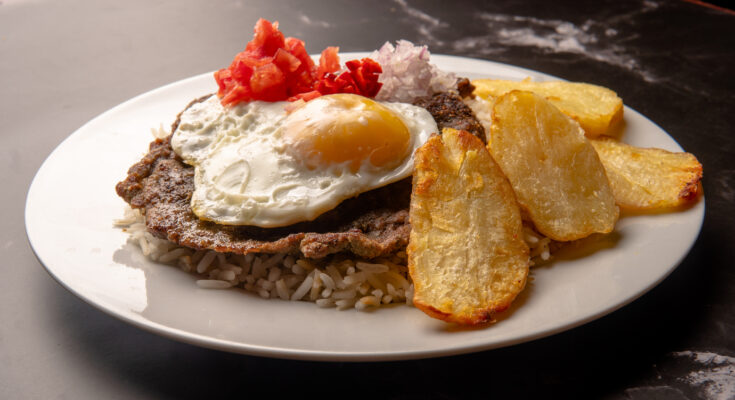 Teller mit Silpancho aus Reis, paniertem Fleisch, Spiegelei, Kartoffeln und Gemüse.