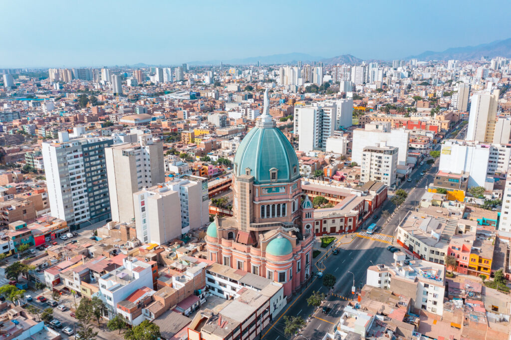 Luftaufnahme von Lima mit einer grün bekuppelten Kirche, dichten Wohnblocks und Bergen am Horizont.