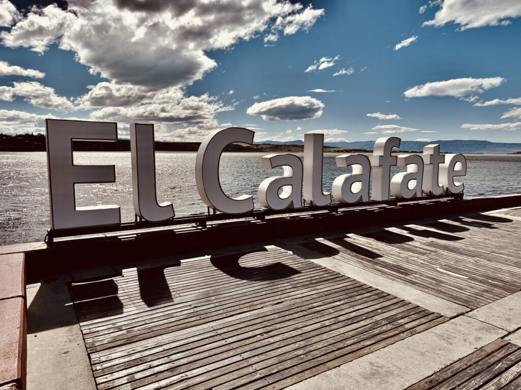 Große „El Calafate“-Buchstaben auf einer Holzpromenade am See unter Wolken.