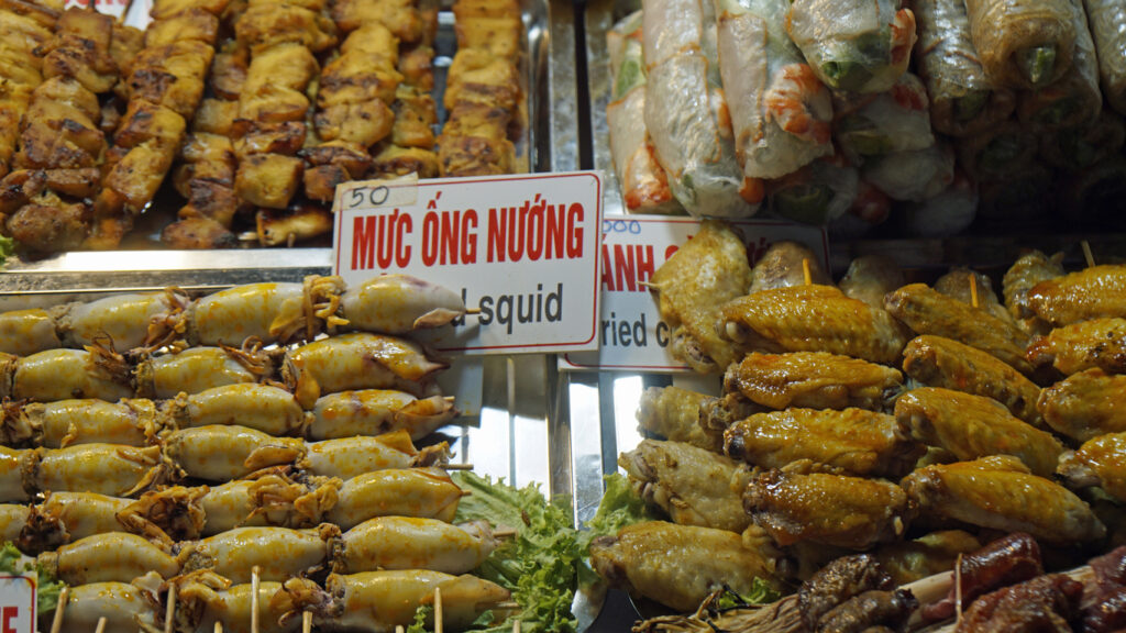 Gegrillter Tintenfisch, Hähnchenflügel und Sommerrollen auf einem Marktstand.