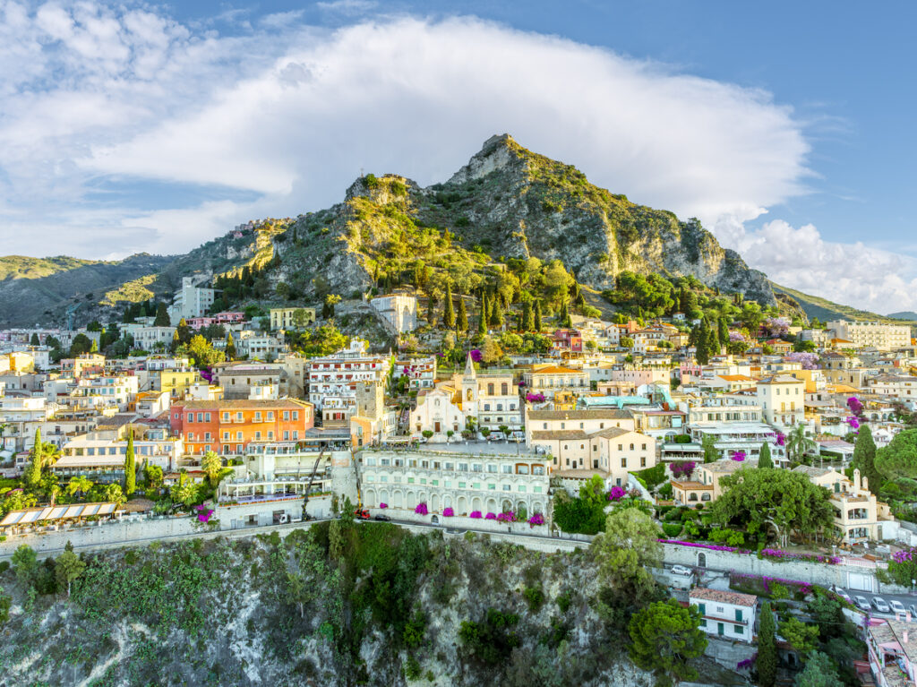 Blick auf Taormina mit dicht bebauten Terrassen, mediterranem Grün und markantem Fels.