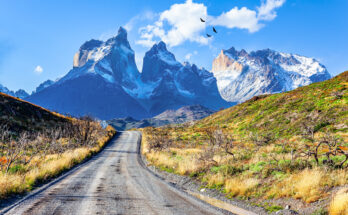 Eine Schotterstraße führt durch Steppe auf die gezackten, verschneiten Berge des Torres del Paine zu.