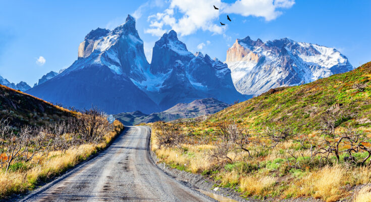 Eine Schotterstraße führt durch Steppe auf die gezackten, verschneiten Berge des Torres del Paine zu.