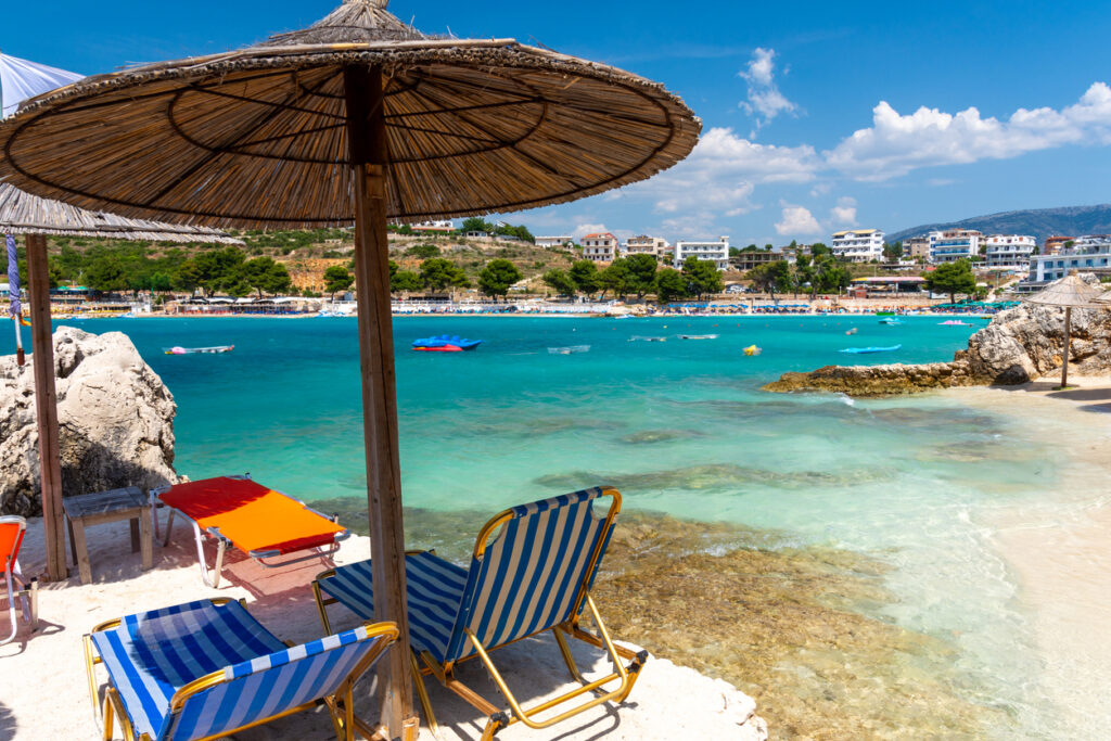 Strandliegen und Strohschirme stehen am hellen Ufer von Ksamil gegenüber der bebauten Bucht.
