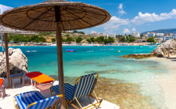 Strandliegen und Strohschirme stehen am hellen Ufer von Ksamil gegenüber der bebauten Bucht.