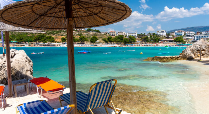 Strandliegen und Strohschirme stehen am hellen Ufer von Ksamil gegenüber der bebauten Bucht.