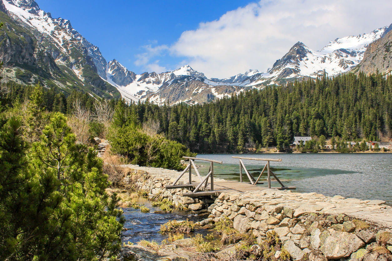 Holzsteg über einem Bach führt zum klaren See vor schneebedeckten Gipfeln.
