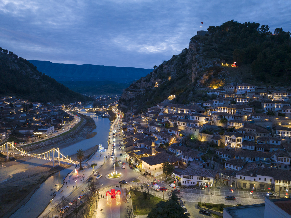 Luftaufnahme der beleuchteten osmanischen Hanghäuser von Berat mit Promenade, Brücken und dem Osum in der Dämmerung.