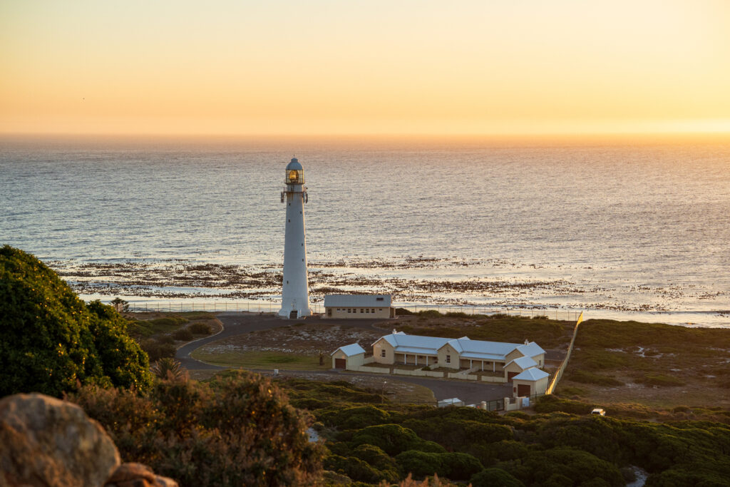 Schlanker weißer Leuchtturm mit Nebengebäuden im warmen Abendlicht.