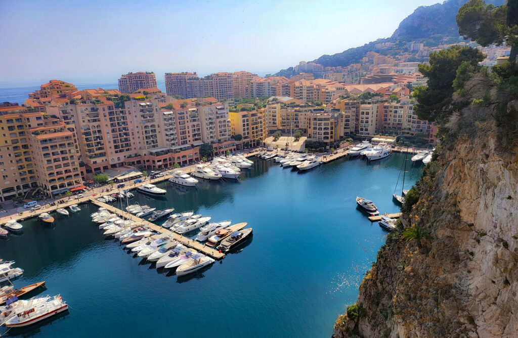 Yachthafen in einer geschützten Bucht unter steilen Felsen mit sandfarbenen Wohnhäusern.